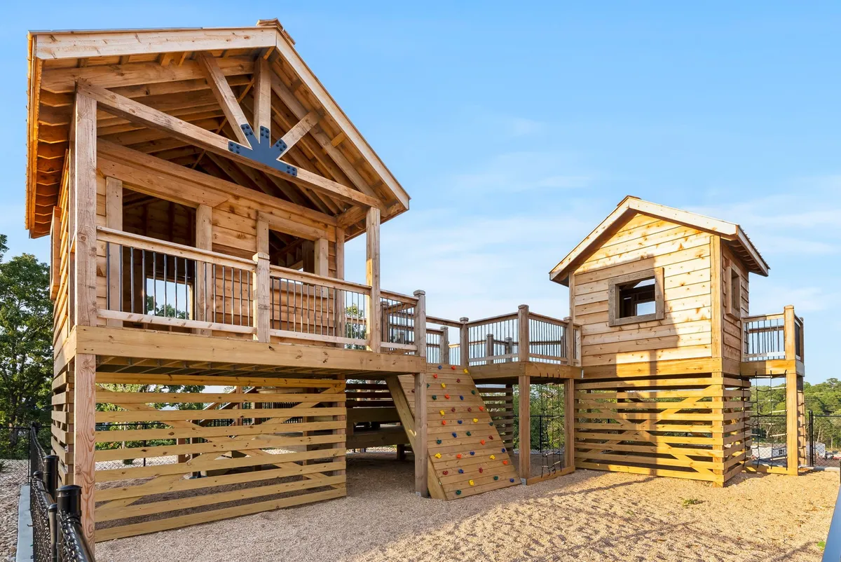 A vibrant exterior playground featuring a wooden playhouse, climbing structure, slide, and staircase. Children are seen playing and exploring, surrounded by greenery that adds to the natural and adventurous setting.