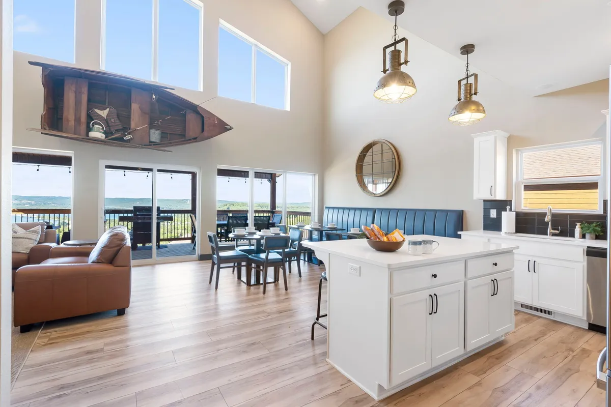 This image showcases a welcoming open-concept living space with large windows offering panoramic views, a modern kitchen island, comfortable seating, and stylish suspended boat decor. The scene is filled with natural light, illustrating togetherness and easy flow between living, kitchen, and dining areas.