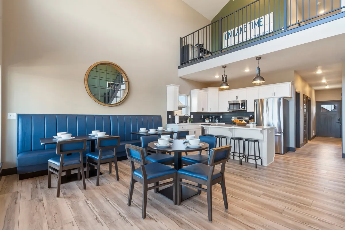 A cheerful, inviting dining area featuring blue upholstered booth seating, a round dining table beautifully set for a meal, and an open kitchen with modern appliances in the background. Sunlight brightens the space, emphasizing communal dining and a homey atmosphere.