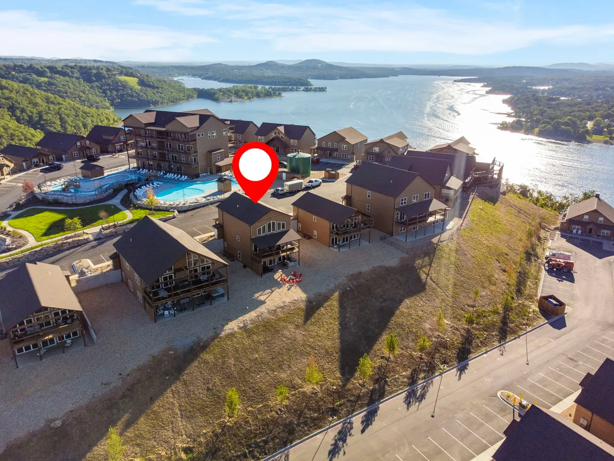 A cheerful exterior scene featuring a swimming pool beside landscaped gardens, with Table Rock Lake shimmering nearby and residential buildings in the background. The sunlit pool area is ready for family fun, barbecues, and easy transitions between poolside lounging and lakeside activities.