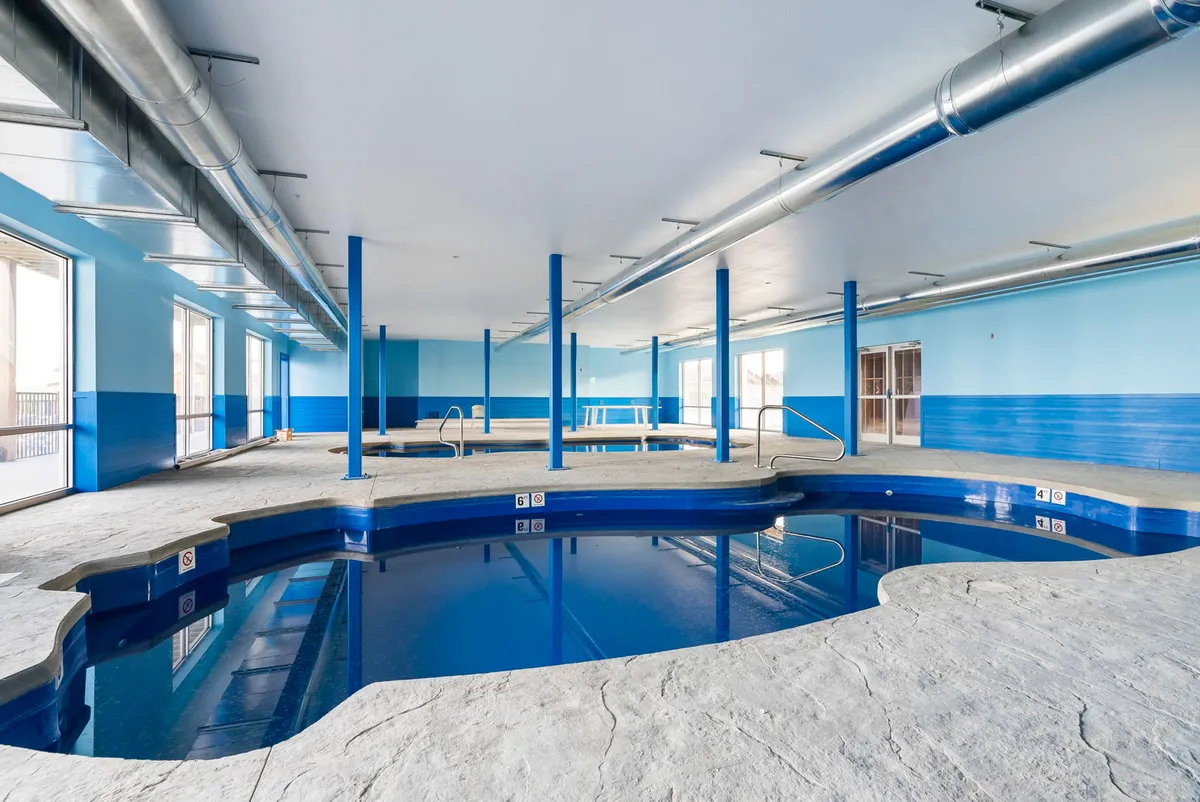 This image showcases a serene indoor pool area with blue walls, large windows letting in natural light, and metallic pipes lending a modern touch. People can be seen swimming and relaxing by the pool, while others are socializing or exercising, capturing the lively and communal atmosphere of the resort's amenities.