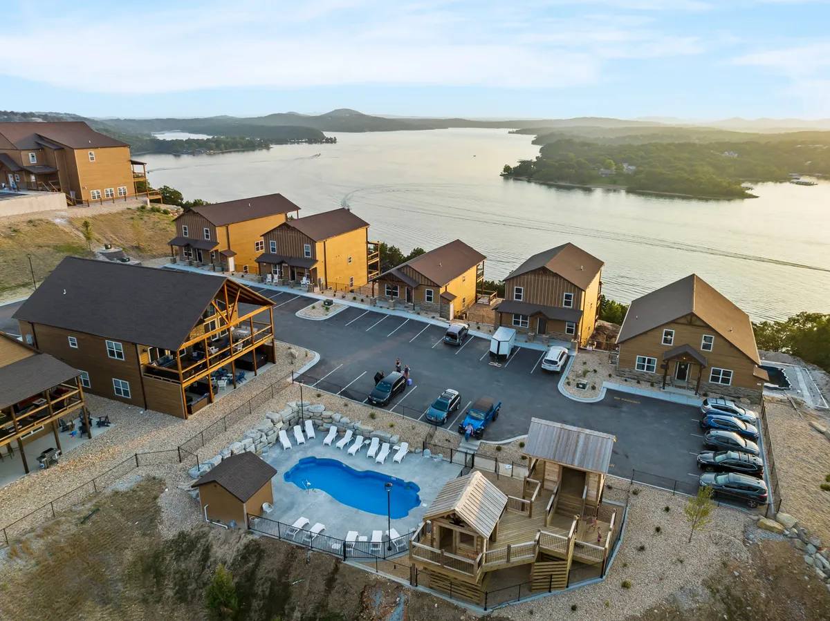 An exterior view of the recreational zone, highlighting the swimming pool, a fun playground structure, a row of cozy cabins, and a scenic lake view in the distance. The environment is vibrant and family-friendly, with children playing, families gathering, and guests relaxing by the pool.