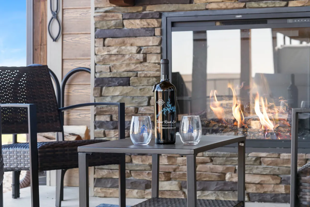 A cozy outdoor firepit area featuring Adirondack chairs arranged around a glowing fire, set against a stone wall and open sky. The setting invites guests to gather and unwind as dusk falls.