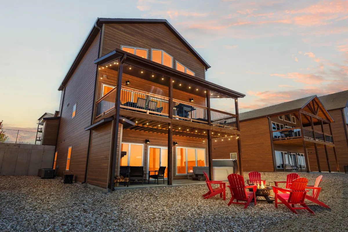 A cozy outdoor gathering area illuminated by string lights, featuring red Adirondack chairs surrounding a fire pit and a balcony above. The setting suggests inviting evenings spent socializing, unwinding, and making memories under the stars.