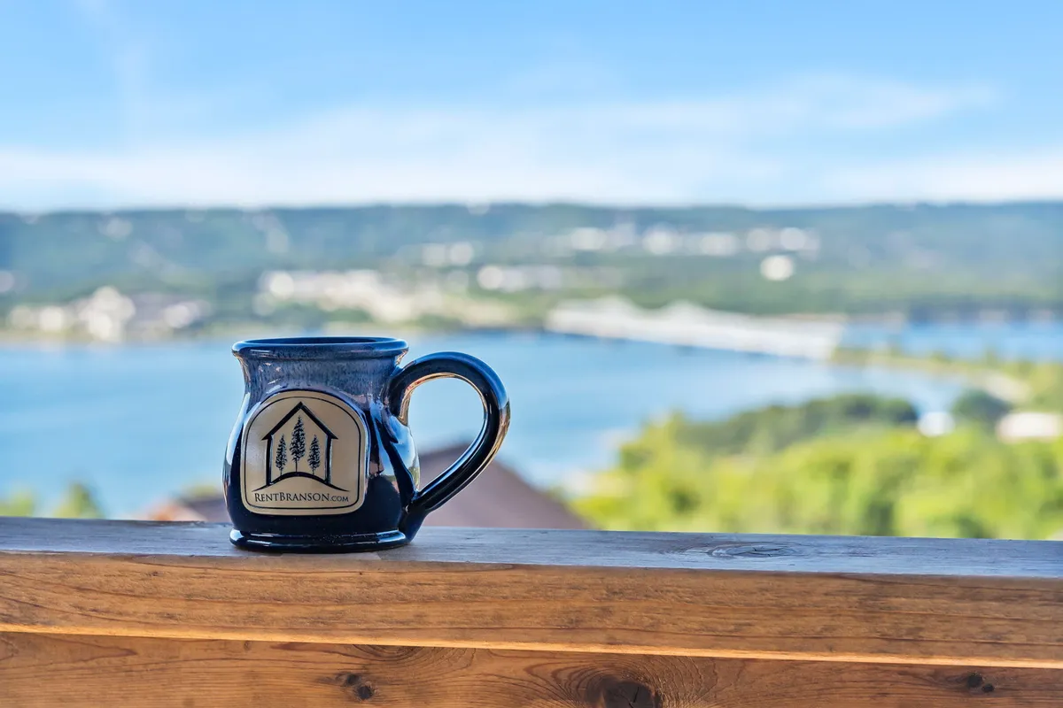 A peaceful scene of a coffee mug resting on a balcony railing, overlooking a lush, detailed landscape with the lake in the distance. The view evokes a sense of calm and connection to nature, ideal for guests savoring quiet moments before or after day trips.