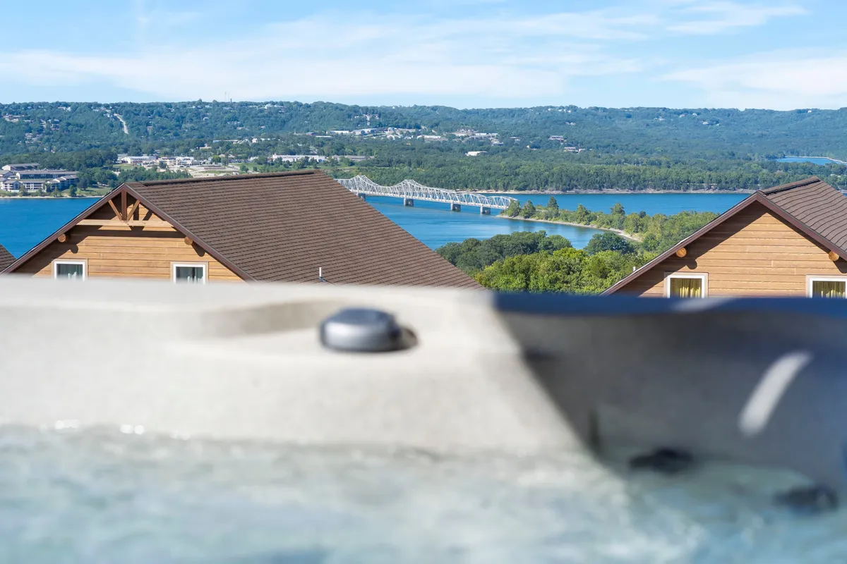 A tranquil outdoor view featuring a jacuzzi on a deck overlooking a scenic lake, with wooden homes and a bridge visible in the distance. The setting is serene, ideal for relaxing after a day of adventure, and highlights the natural beauty of the Ozarks.