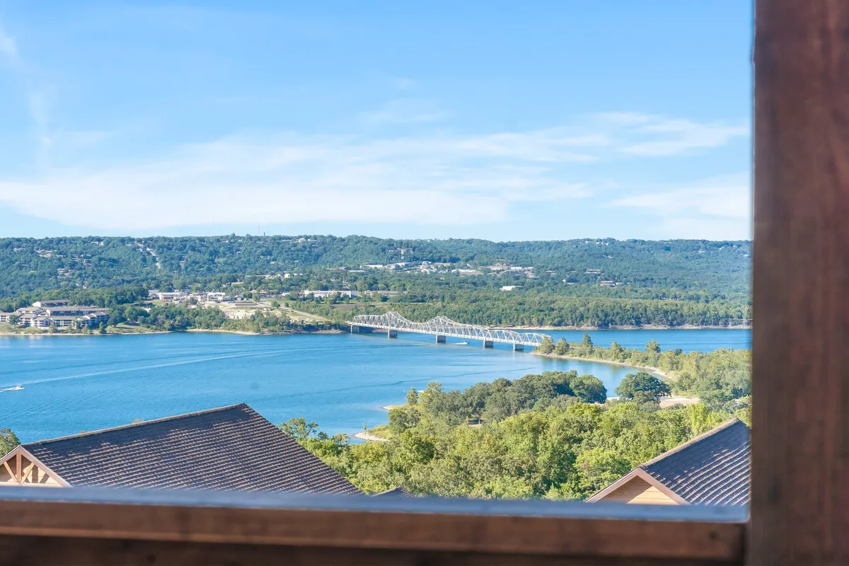 A sweeping, tranquil view of Table Rock Lake with a prominent bridge in the mid-ground, rolling hills and lush trees along the water’s edge. The glassy lake surface is dotted with occasional boats, and the sky glows with sunset reflections, hinting at the area’s calm and beauty.