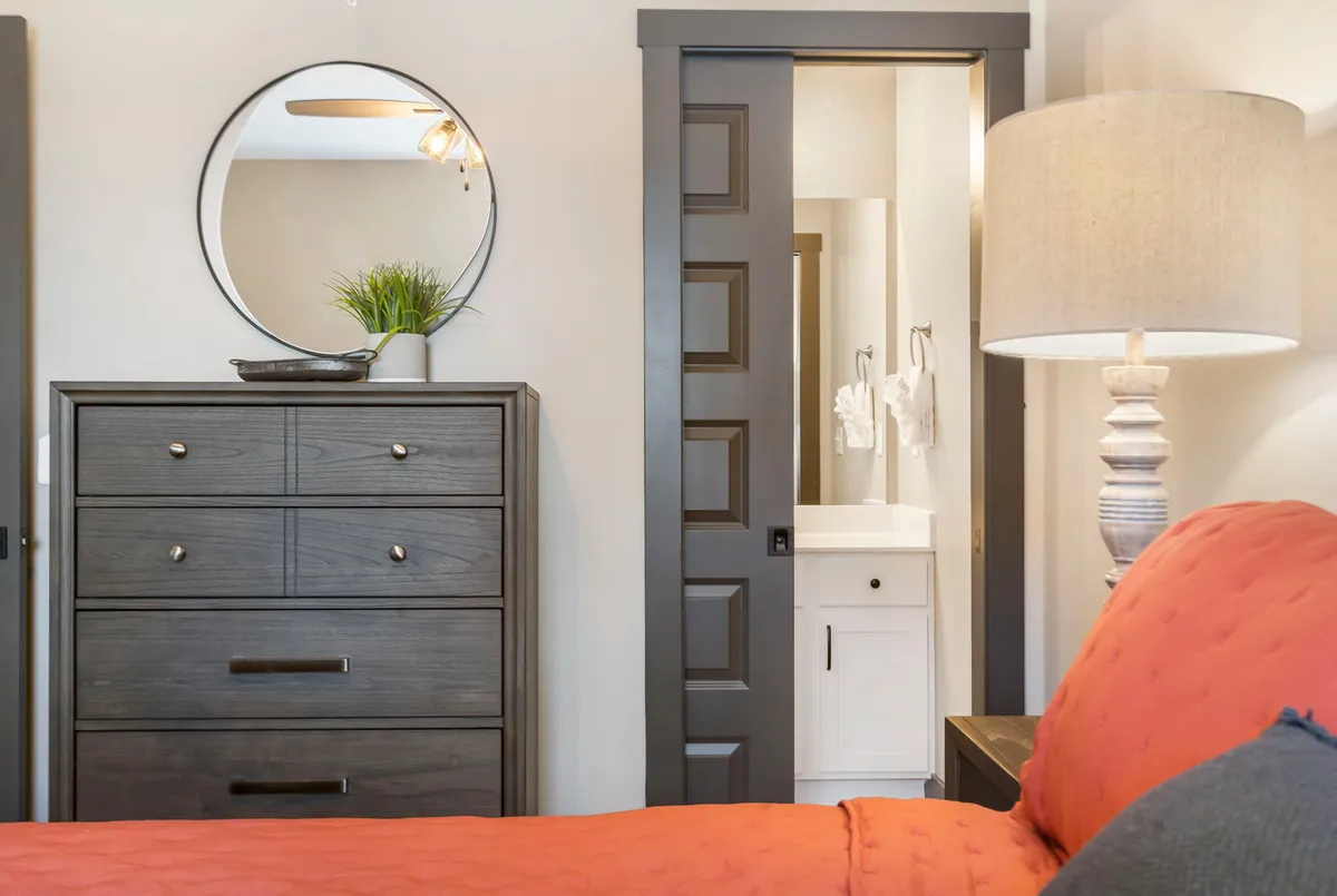 A stylish king bedroom featuring a bed with vibrant orange bedding, a dark dresser, a round mirror, and a table lamp. The space exudes comfort, with an inviting atmosphere ideal for relaxing, reading, or preparing for a restful night.