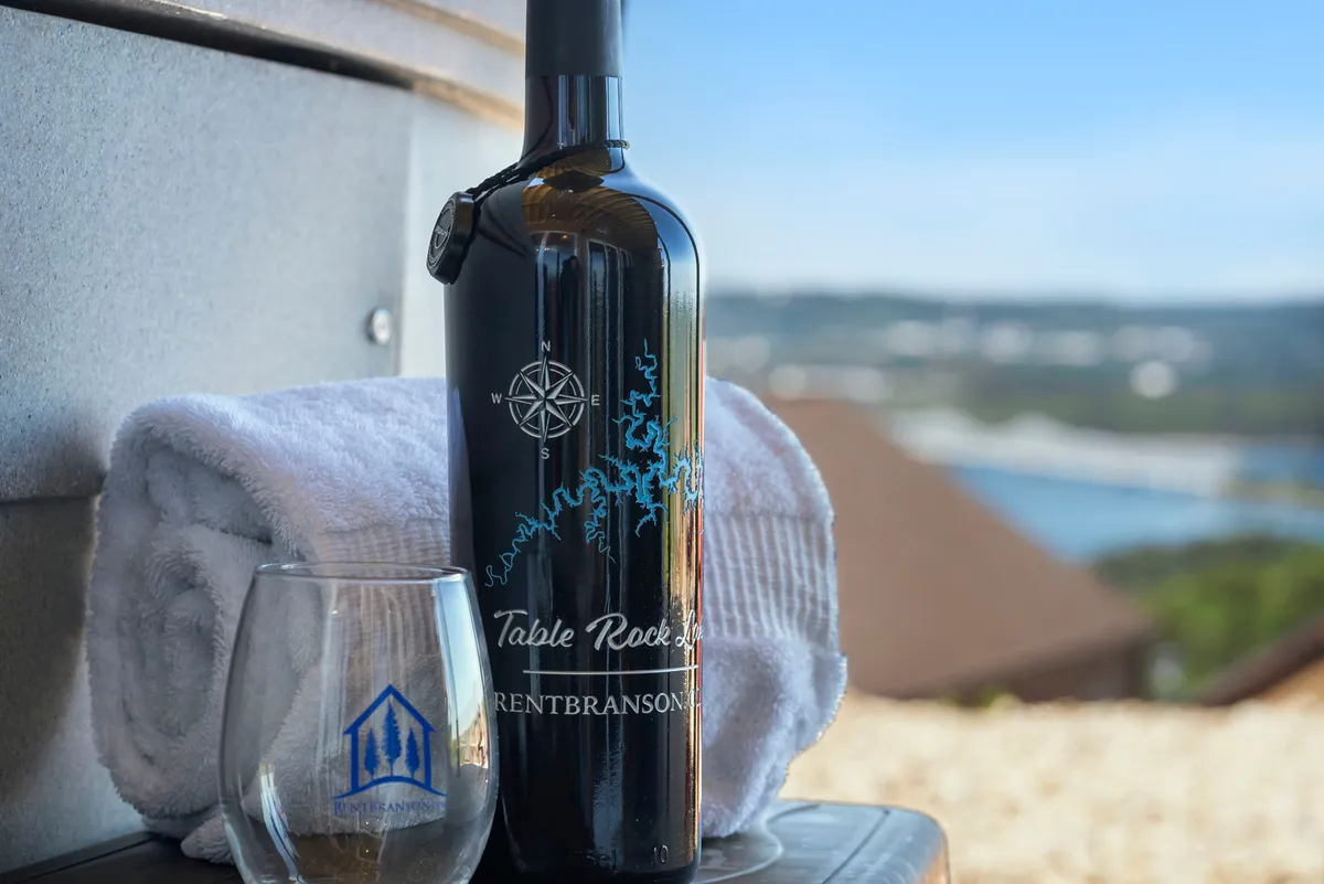 This exterior image captures a wine bottle and glass set on a ledge with a scenic natural background, suggesting a tranquil spot to unwind while admiring the view. The mood is serene and luxurious, perfect for moments of reflection or celebration against the beauty of the Ozarks.