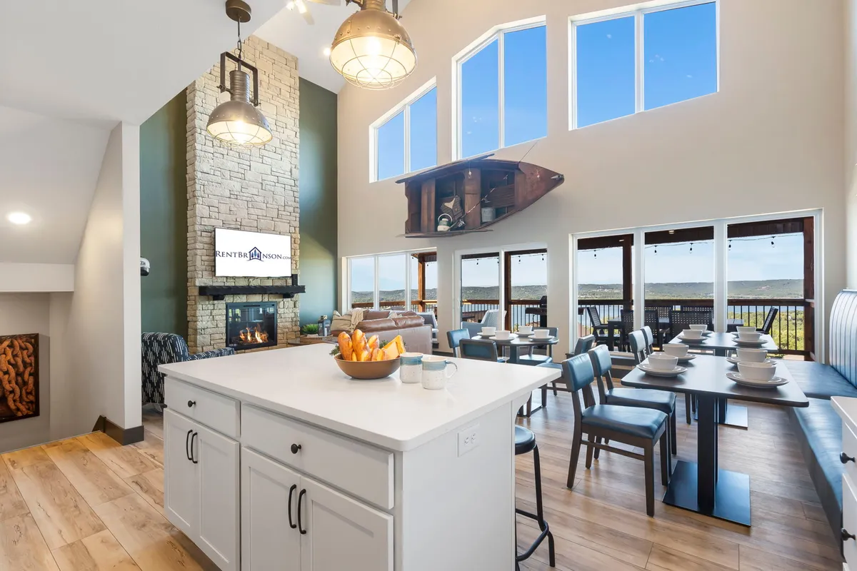 The image showcases a warm and inviting open-concept living space with a grand stone fireplace as the focal point. Expansive floor-to-ceiling windows frame stunning lake views and fill the room with natural light. The spacious kitchen island is set with snacks, and a large dining area is prepared for gathering and meals, all set against the dramatic backdrop of Table Rock Lake and the Kimberling City Bridge.