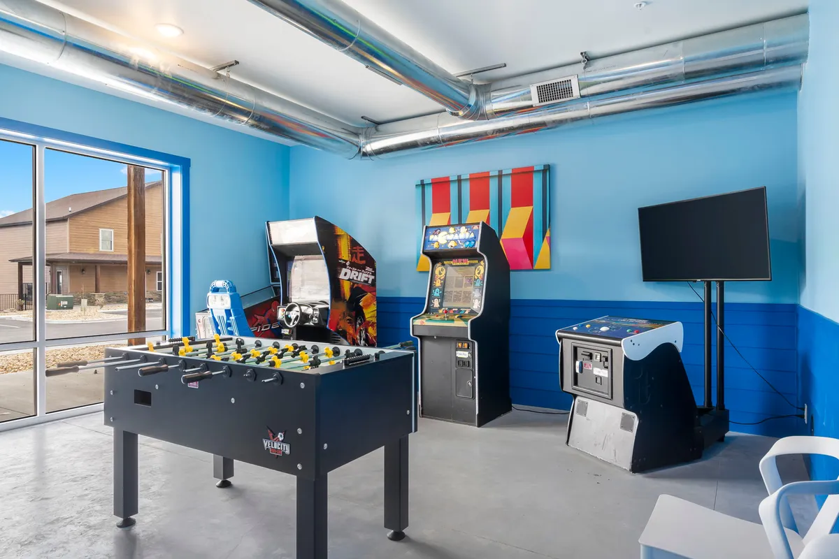 This interior image features a lively game room equipped with a foosball table, arcade machines, a TV screen, and vibrant wall art. The setup is designed for group entertainment and friendly competition, making it an inviting space for all ages to gather and play together.