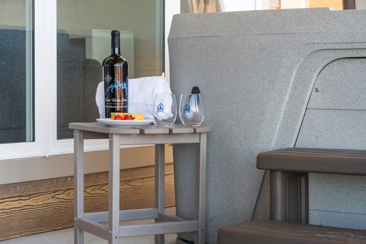 The scene features a close-up of a wine bottle, glasses, fruit platter, and a towel next to the hot tub, emphasizing personal indulgence. The backdrop suggests a peaceful and inviting environment ideal for shared moments, laughter, and relaxation after a day of adventure.